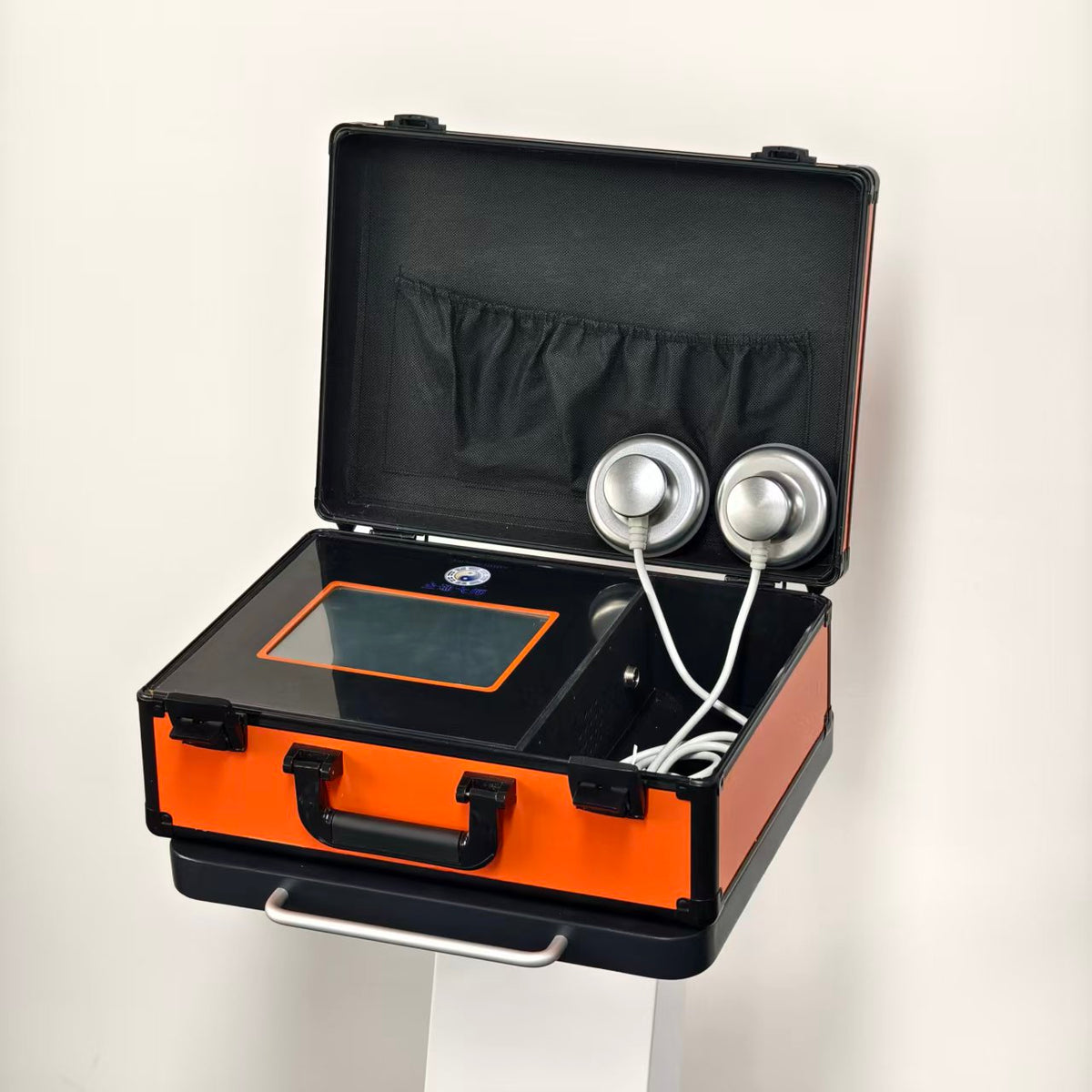 Open orange carry case seen from above at an angle: touchscreen panel on the left, cable compartment on the right, two round stainless-steel massage heads